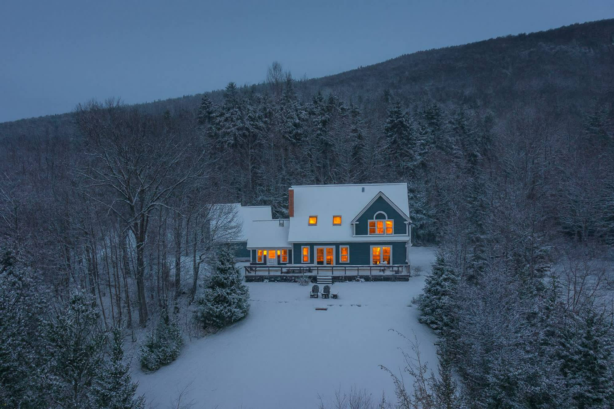 The main house, a blue two-story structure, is covered in fresh snow at dusk, with warm lights glowing from its windows. It's surrounded by snow-dusted evergreen trees and a snowy landscape.