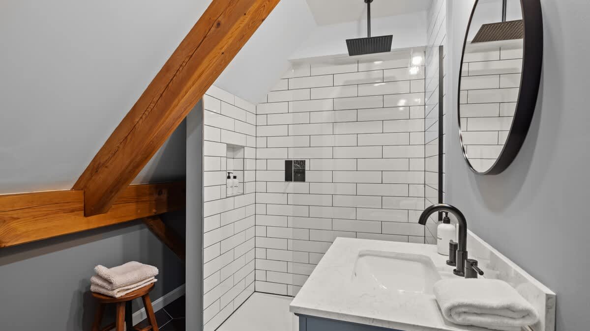 Modern bathroom with a white subway tile shower, black fixtures, a white vanity with a round mirror, and a sloped ceiling with exposed wooden beams.