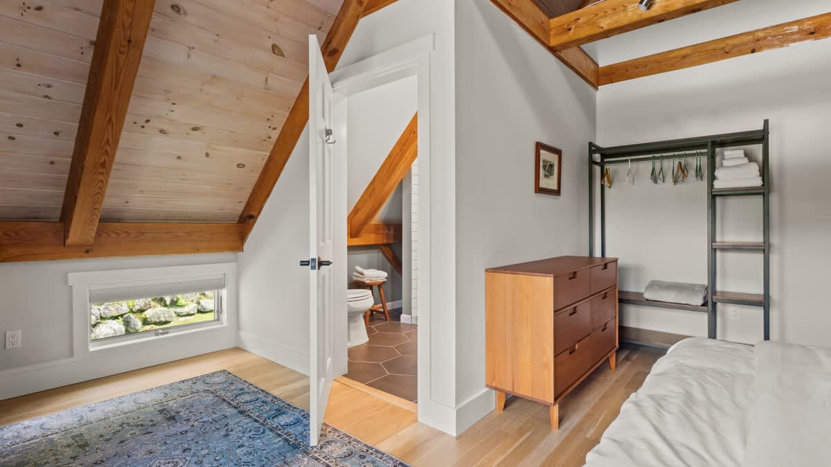 Attic bedroom with exposed wooden beams, light wood floors, and a blue patterned rug, showing a white bed and an open door leading to a bathroom with hexagonal floor tiles.