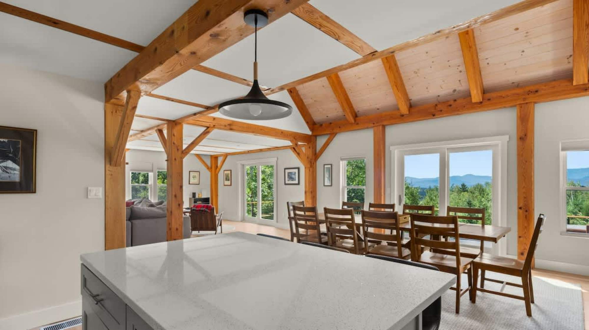 Open-concept dining area with a long wooden dining table and eight matching chairs, featuring exposed wooden beams, hardwood floors, and large windows offering mountain views.