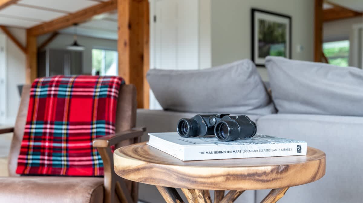 A close-up of a rustic wooden side table in the living room, adorned with a pair of black binoculars and a book, next to a leather armchair draped with a red plaid blanket.