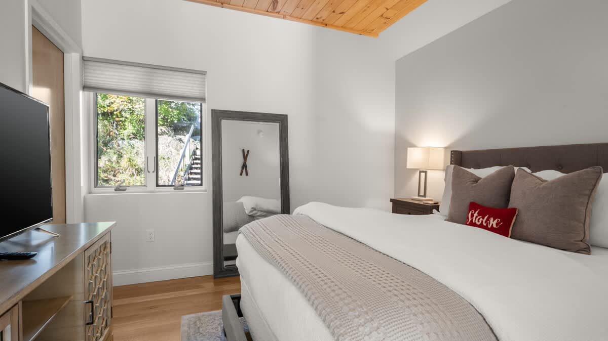 Cozy bedroom featuring a large bed with a grey upholstered headboard and a red 'Stowe' pillow, light wood floors, a window with blinds, and a full-length mirror reflecting the room.