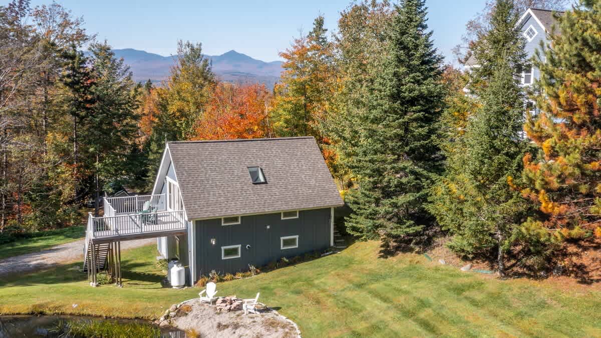 A grey A-frame style guest house with a raised deck overlooks a small pond, surrounded by green grass and autumn trees. Distant mountains are visible under a bright blue sky.
