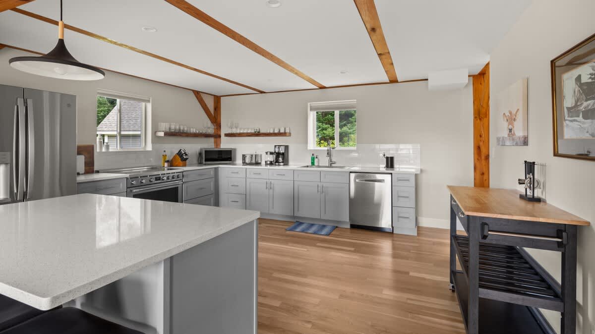 A spacious, modern kitchen with light gray cabinetry, white quartz countertops, stainless steel appliances, and light hardwood floors, accented by exposed wooden beams on the ceiling.