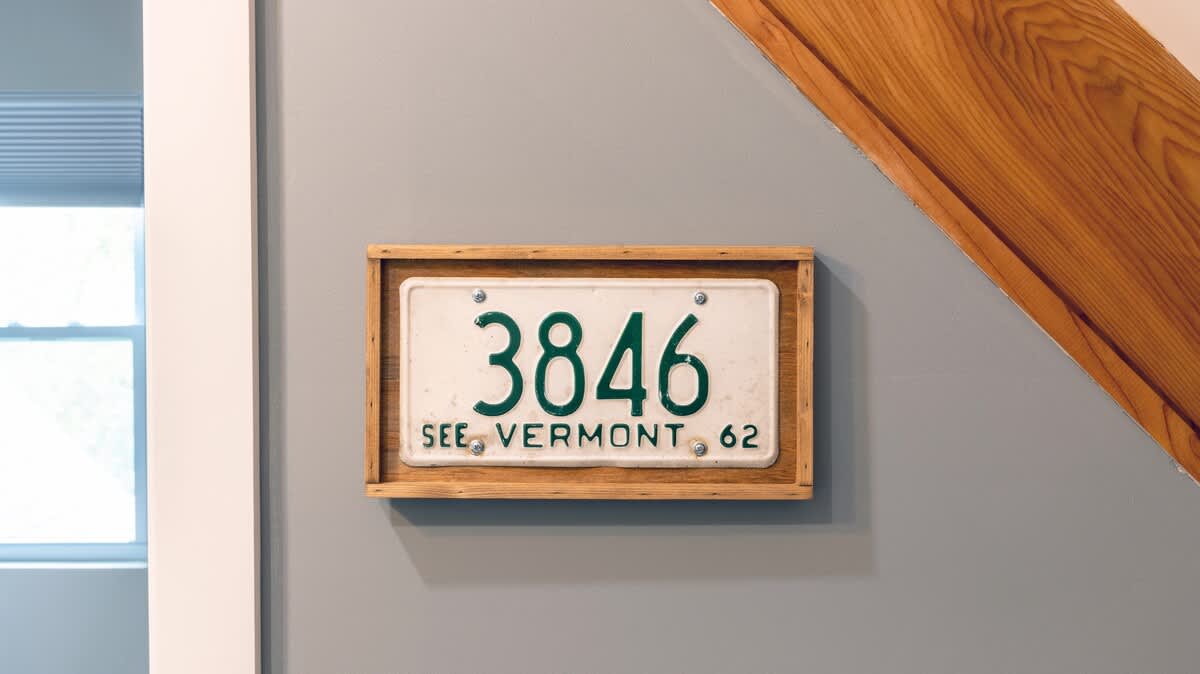 Close-up of a vintage 'SEE VERMONT 62' license plate framed in wood and mounted on a light gray wall, with a wooden beam visible in the background.