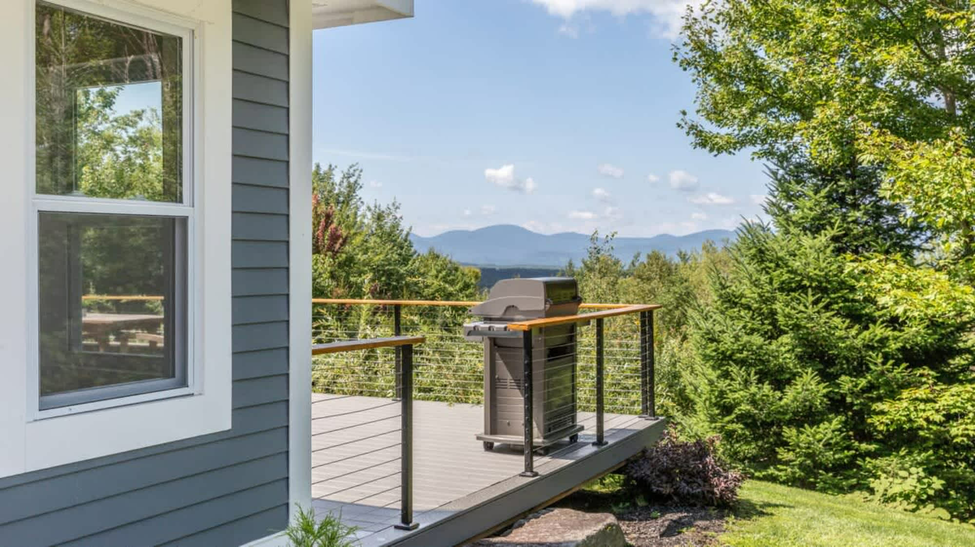 Corner of an outdoor deck with gray composite decking and a black gas grill, offering scenic views of distant mountains and dense green forests under a blue sky.