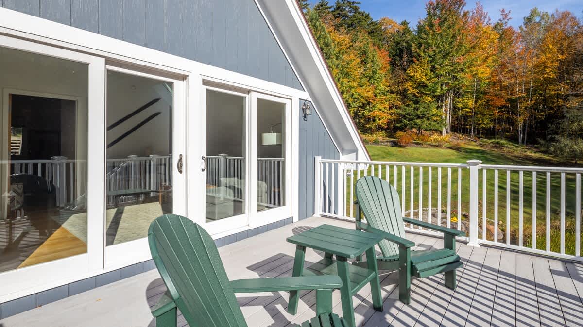 A spacious wooden deck with white railings features two green Adirondack chairs and a matching side table, offering views of a grassy hillside and colorful autumn trees. Large glass sliding doors lead into the house.