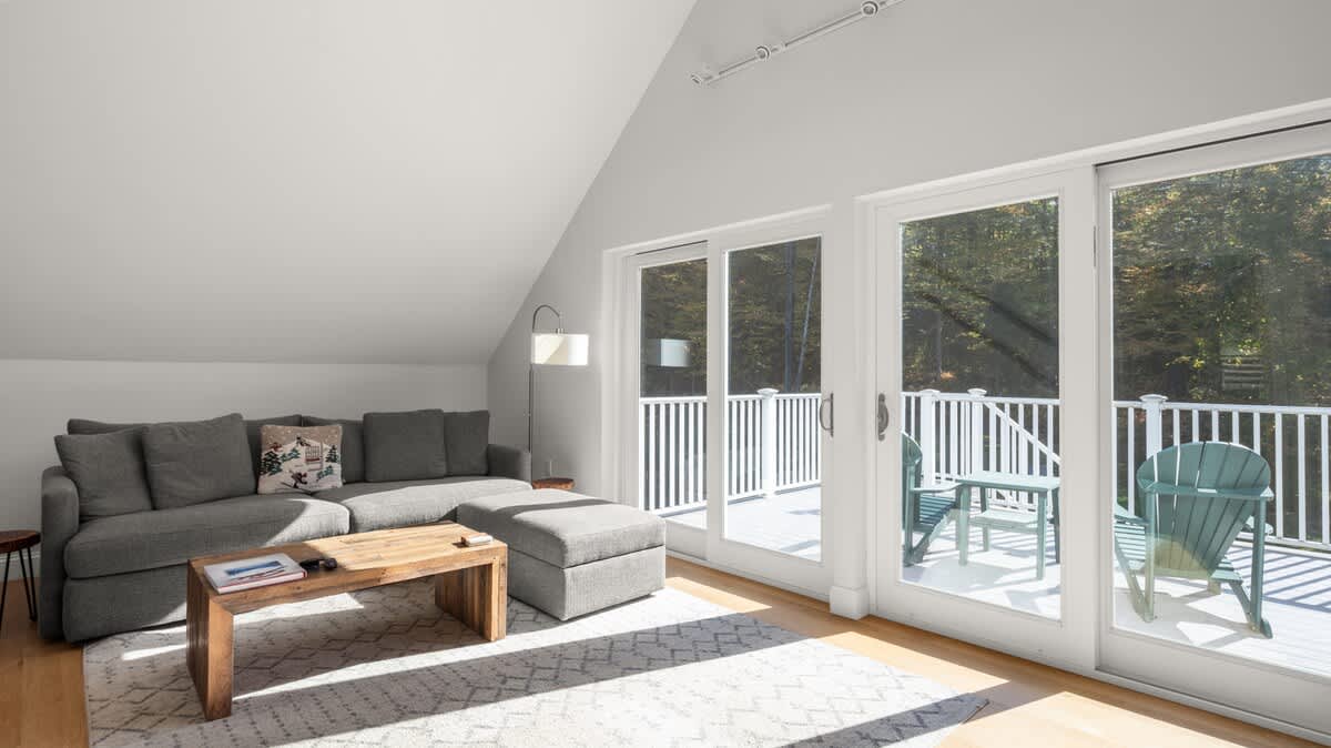 Bright living room with vaulted ceilings, light hardwood floors, and a large gray sectional sofa. Sliding glass doors open to a balcony, allowing ample natural light and views of the surrounding autumn trees.