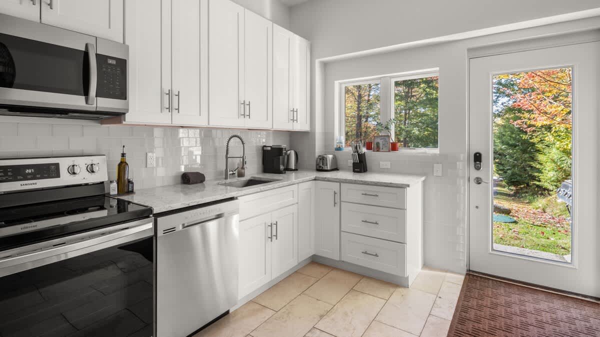 Modern kitchen featuring white shaker cabinets, light granite countertops, white subway tile backsplash, and stainless steel appliances including a microwave, range, and dishwasher. A window and glass-paned door provide natural light and views of the outdoors.