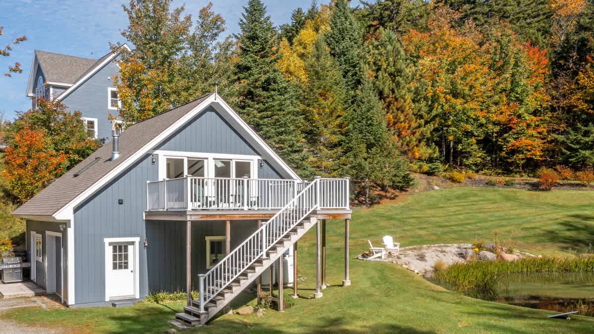 Aerial view of a blue-sided house with a large deck and outdoor stairs, nestled among vibrant autumn trees and overlooking a small pond and grassy yard.