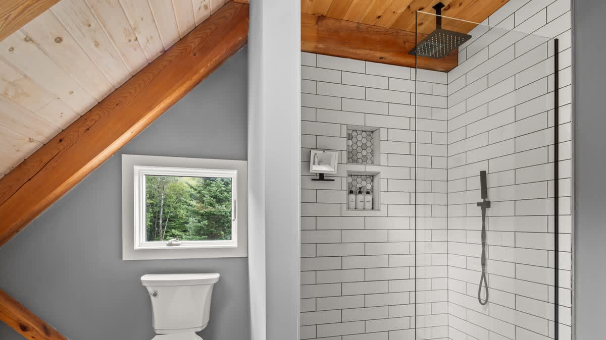 This bathroom features a spacious shower with white subway tiles and a black rain shower head, adjacent to a toilet positioned beneath a window. The room maintains the home's aesthetic with a slanted ceiling and exposed wooden beams.