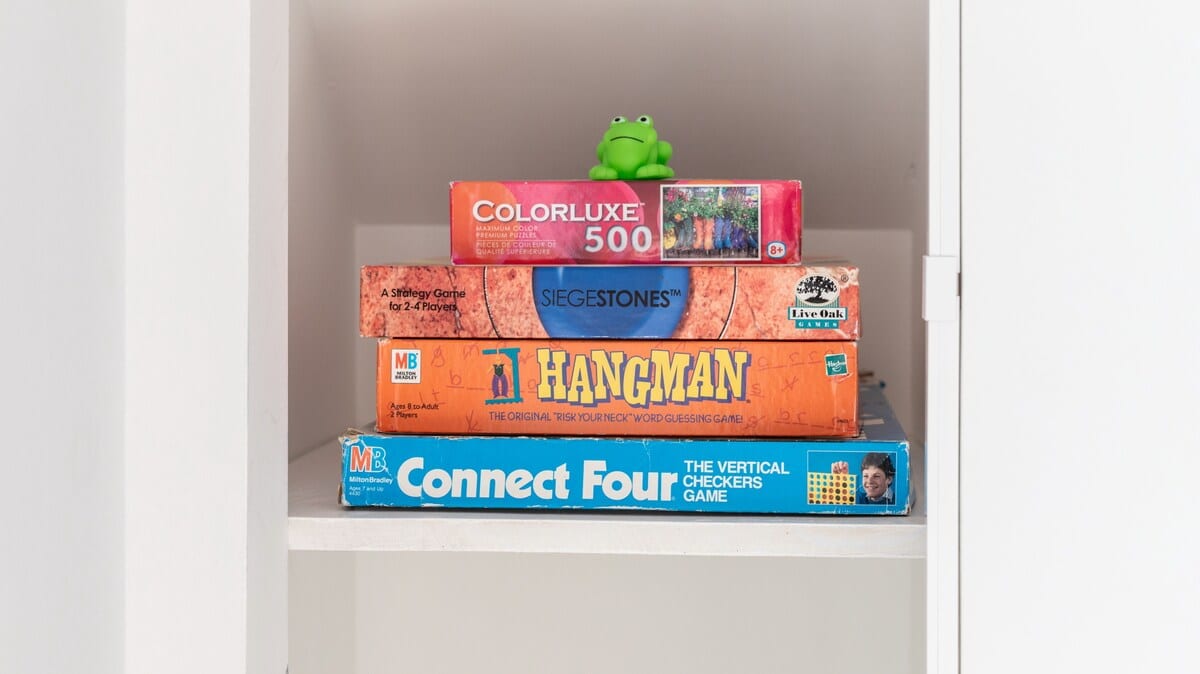 A stack of classic board games including Colorlux 500, Siegestones, Hangman, and Connect Four, neatly stored in a white cabinet, topped with a small green frog figurine.