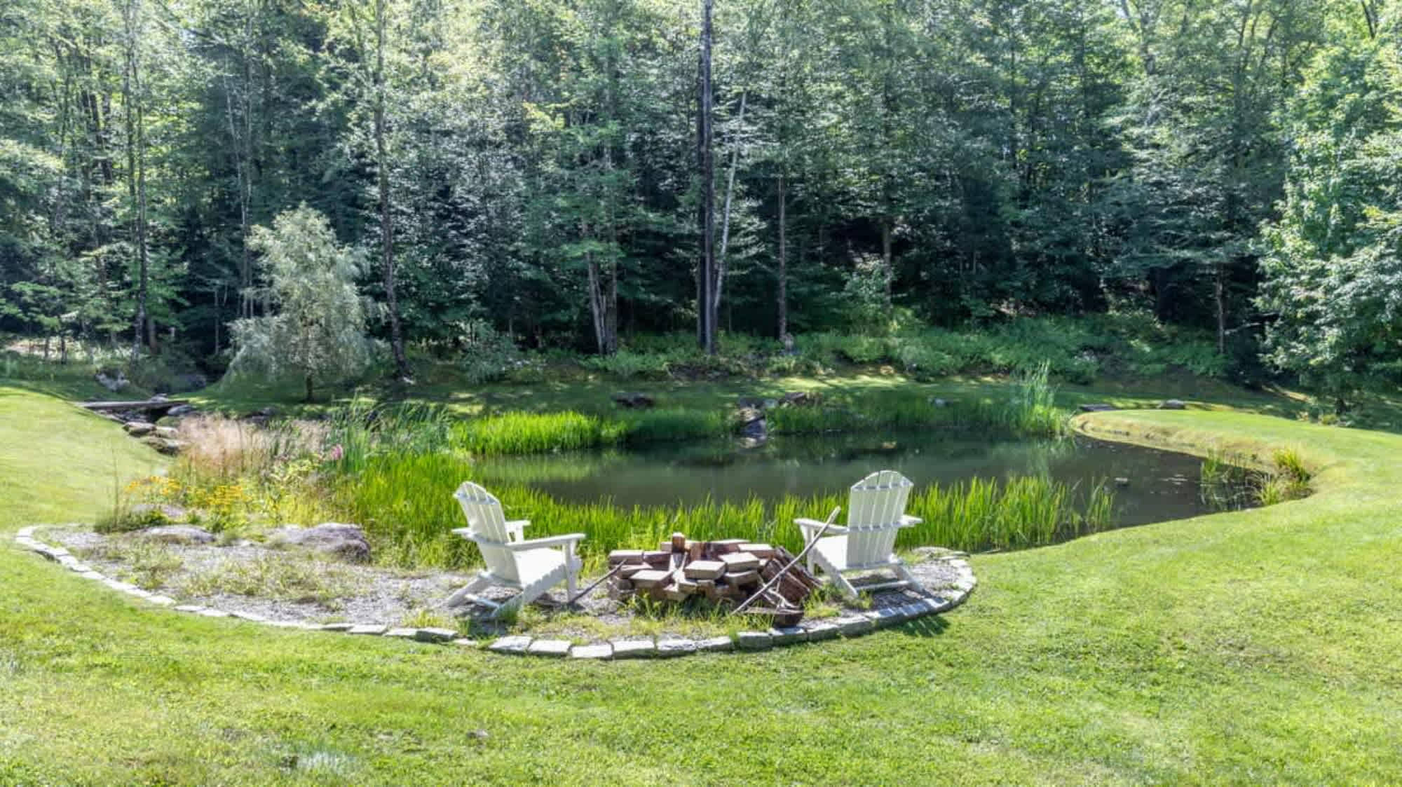 Serene backyard area with a stone-ringed fire pit and two white Adirondack chairs, overlooking a tranquil pond surrounded by lush green grass and dense forest.