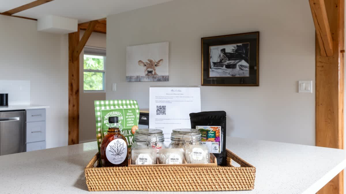 A kitchen counter with a welcome tray featuring local maple syrup, various teas in glass jars, and a printed welcome note with a QR code, set against a light gray wall with exposed wooden beams.