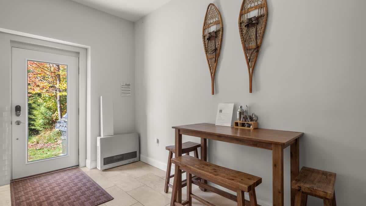 Bright entryway with light gray walls, tiled floor, a small wooden dining table with benches, and decorative snowshoes mounted on the wall. A glass-paned door offers a view of colorful autumn trees outside.