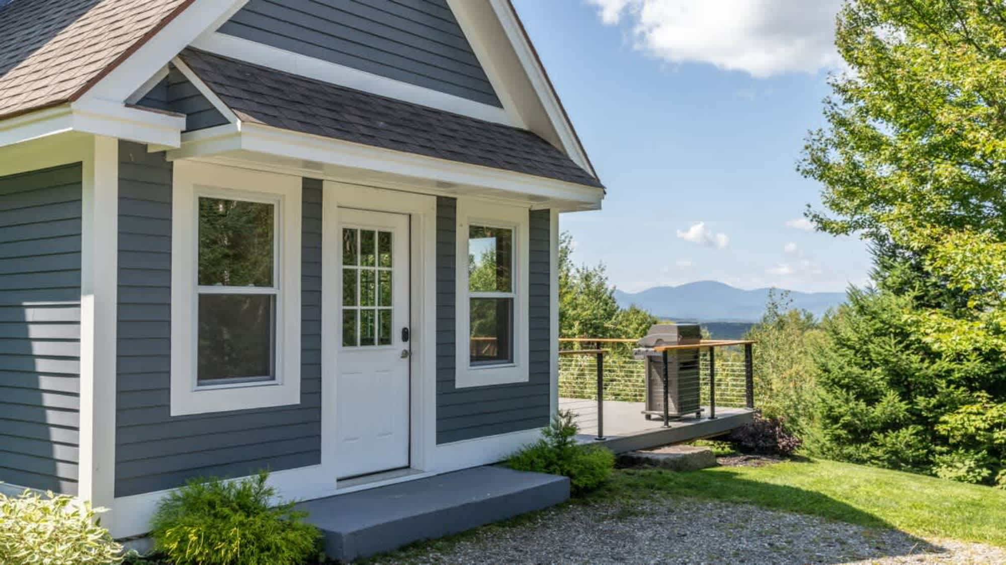 A small, modern cottage with dark gray siding and white trim, featuring a white front door with glass panes and a small deck with a grill, offering distant mountain views.