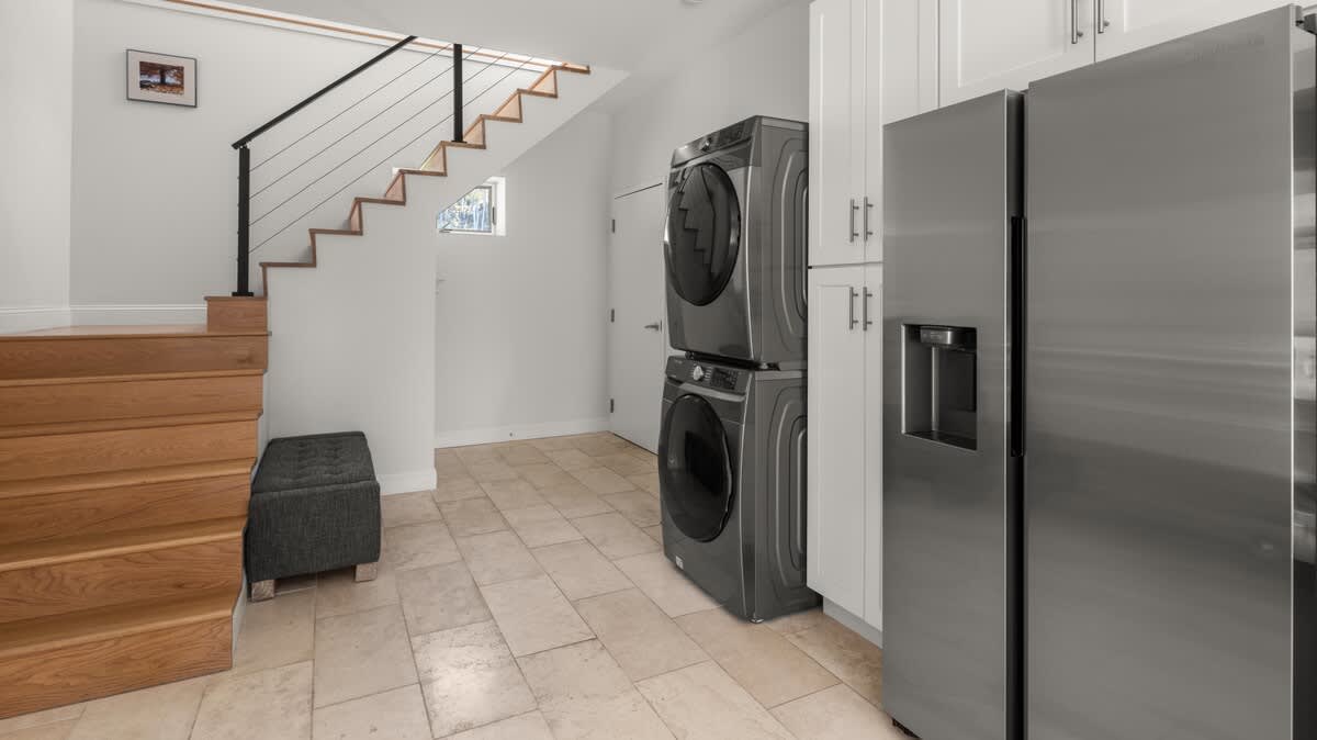 Laundry area with stacked stainless steel washer and dryer units, a large stainless steel refrigerator, and white cabinets, located next to a wooden staircase with black railings.