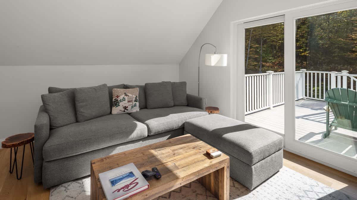 Spacious living room with light hardwood floors and a large gray sectional sofa, centered around a rustic wooden coffee table. Sliding glass doors lead to an outdoor balcony with white railings and green Adirondack chairs.