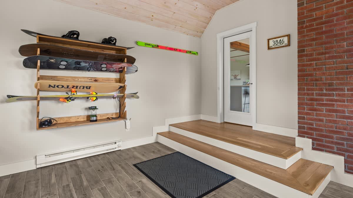 A functional mudroom area with wood-look tile flooring, a wooden ski and snowboard rack, and a small set of wooden stairs leading to a white door with a glass panel. A brick accent wall adds warmth.