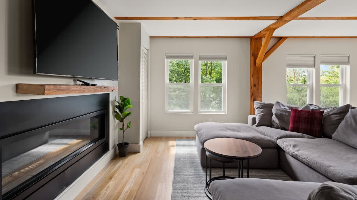 A cozy living room features a large grey sectional sofa and a modern fireplace with a wall-mounted TV above. Hardwood floors and exposed wooden beams complement the bright space, with windows offering views of green trees.
