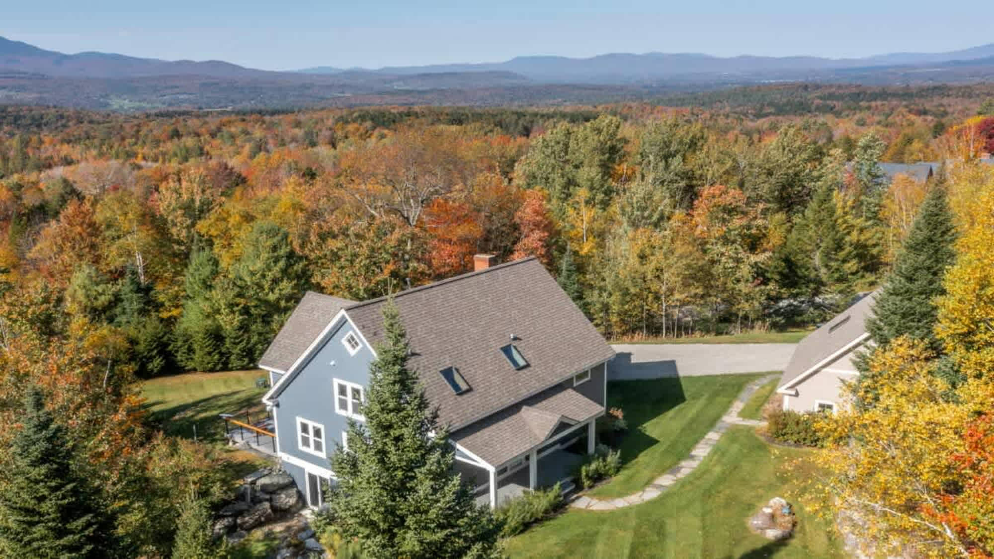 Aerial view of the blue house with a gray roof, surrounded by vibrant autumn trees and a lush green lawn, with distant mountains visible under a clear blue sky. A second smaller building is also visible.