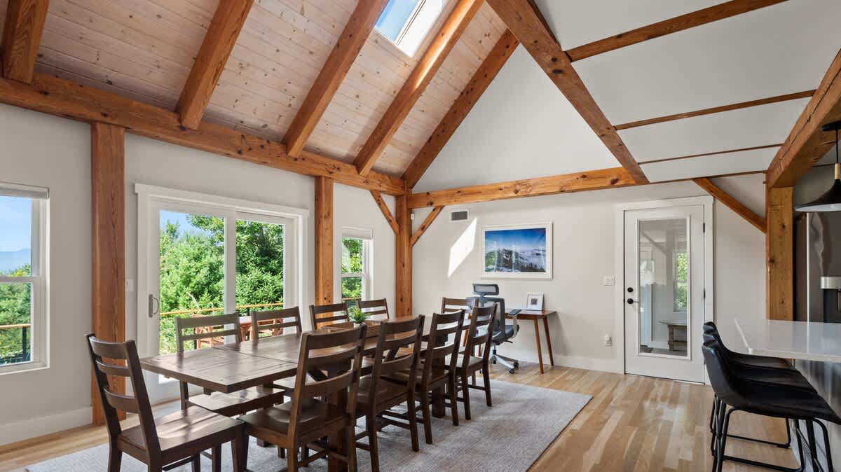 A bright dining area features a long wooden table with seating for ten, set under a vaulted ceiling with exposed timber beams and a skylight. Large windows and a sliding door offer views of lush greenery, with a small office nook visible in the background.