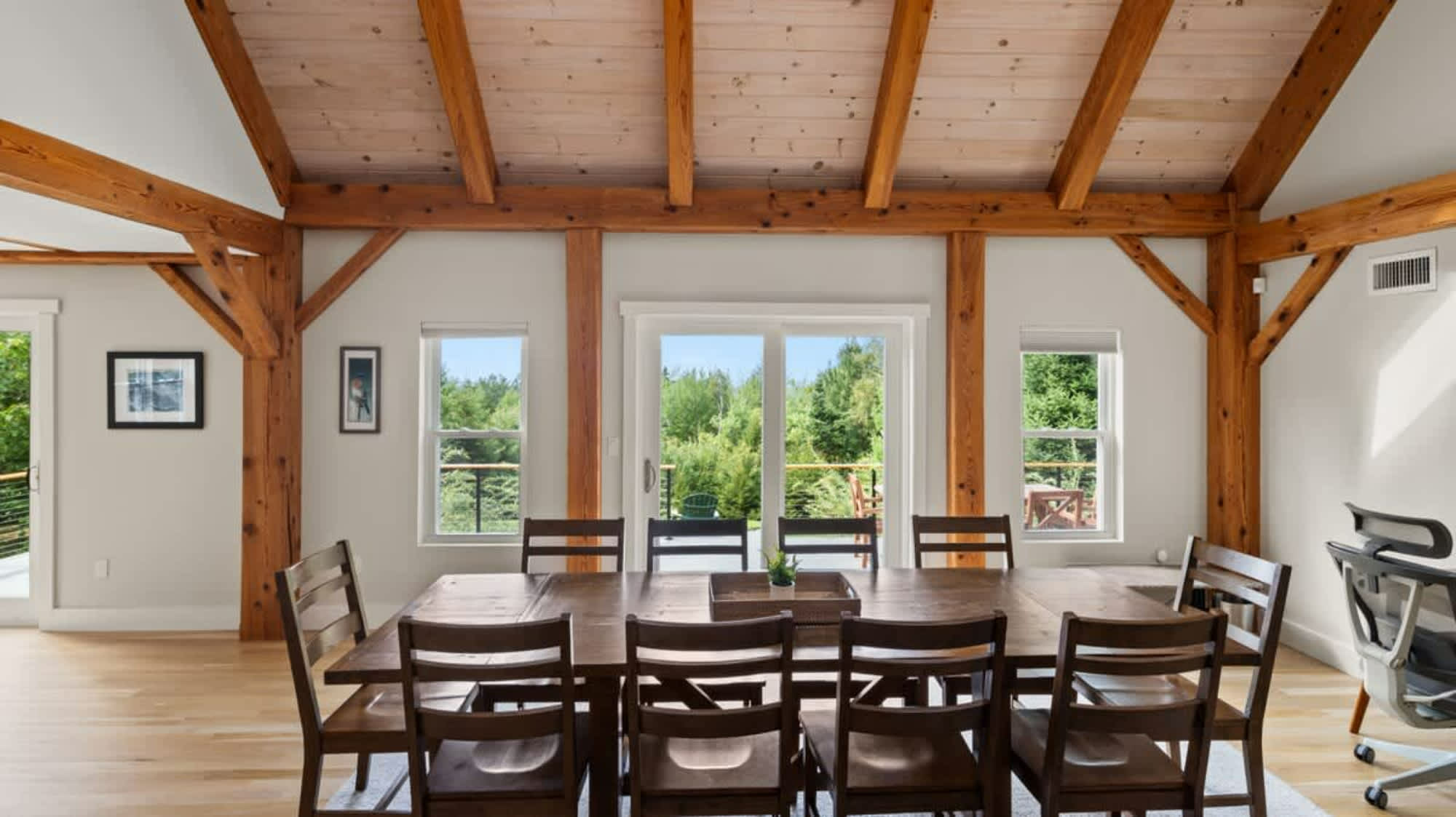 Spacious dining area with a large wooden table and chairs, featuring a vaulted ceiling with exposed wooden beams, light walls, and large windows and a sliding door leading to an outdoor space.