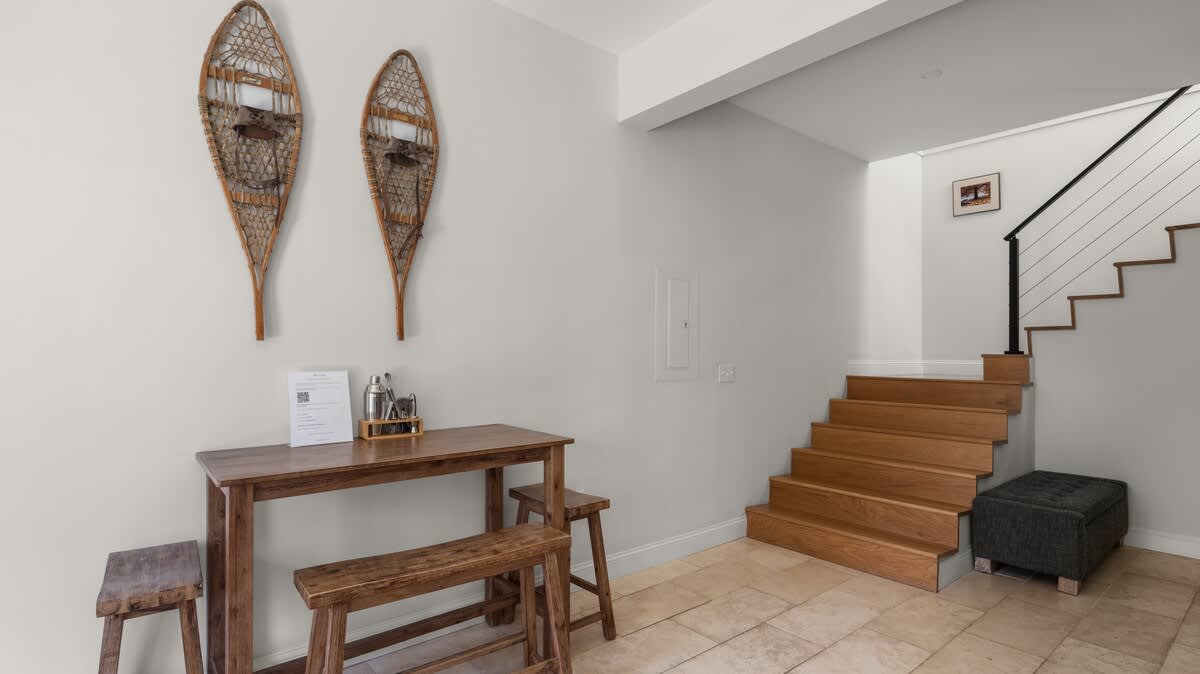 Entryway area with light gray walls, tiled floor, a small wooden table with benches, and decorative snowshoes, leading to a modern wooden staircase with black metal railings.