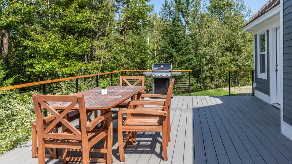 Outdoor deck with gray composite decking, a wooden dining table, and chairs, featuring a black gas grill and surrounded by lush green trees.