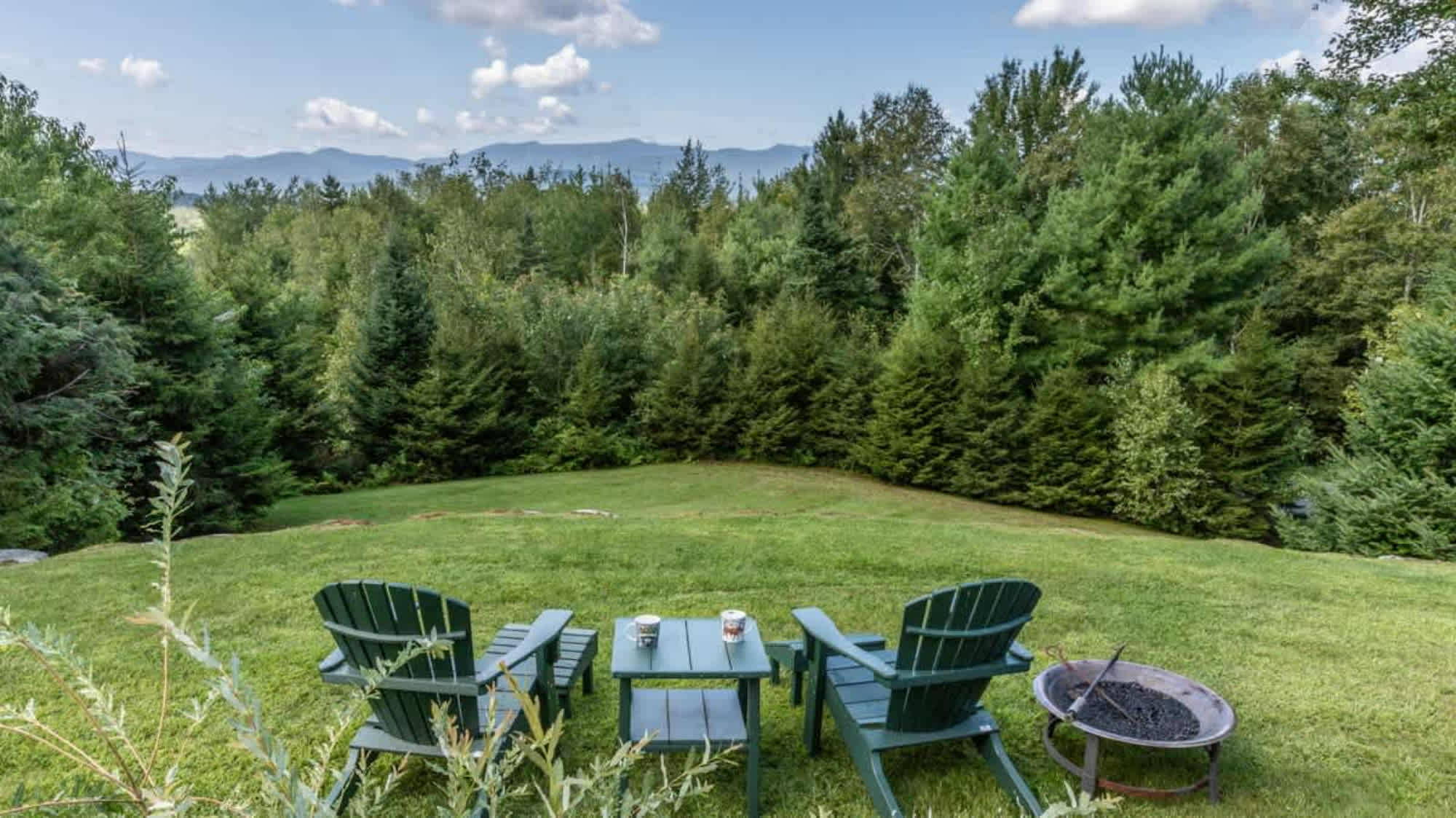 Lush green lawn with two dark green Adirondack chairs and a matching side table, facing a dense forest and distant mountains under a partly cloudy sky, with a metal fire pit to the right.