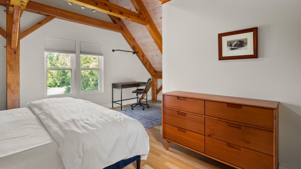 Bright attic bedroom with a white bed, light wood dresser, and a dark desk, featuring exposed wooden beams, a sloped ceiling, and large windows overlooking lush greenery.