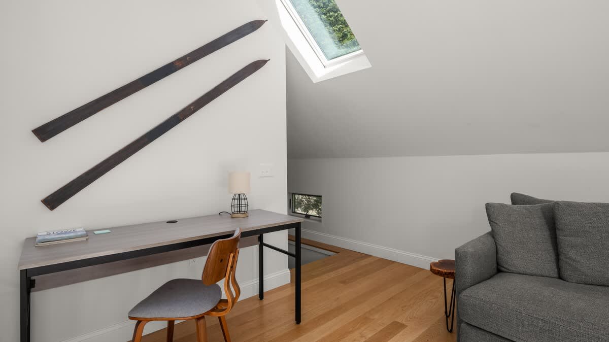 A bright attic workspace features a light wood desk and chair, a gray sofa, and two vintage skis mounted on the white sloped wall. A skylight and small window provide natural light, illuminating the light hardwood floor.