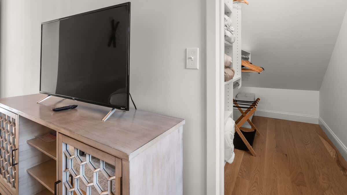 A flat-screen TV rests on a light wood media console with decorative doors, positioned next to an open closet revealing shelving and hangers. The closet offers organized storage for clothing and linens.