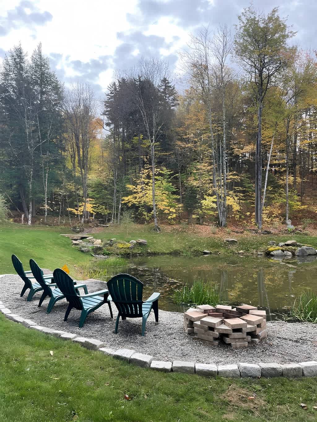 A cozy fire pit area features three green Adirondack chairs arranged around a stone paver fire pit, set beside a tranquil pond. The scene is backed by a forest of trees displaying vibrant autumn foliage under a cloudy sky.