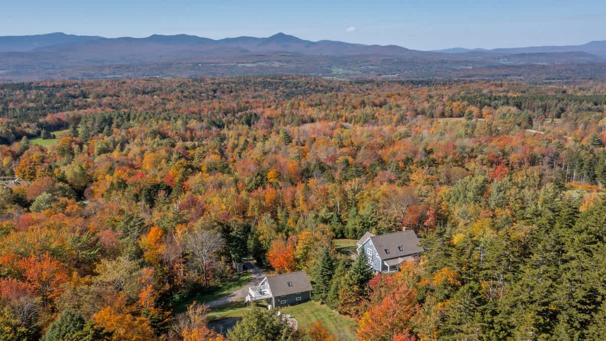 A stunning aerial view showcases two charming houses nestled amidst a vast expanse of vibrant autumn forest, with rolling mountains visible in the distant background under a clear sky.