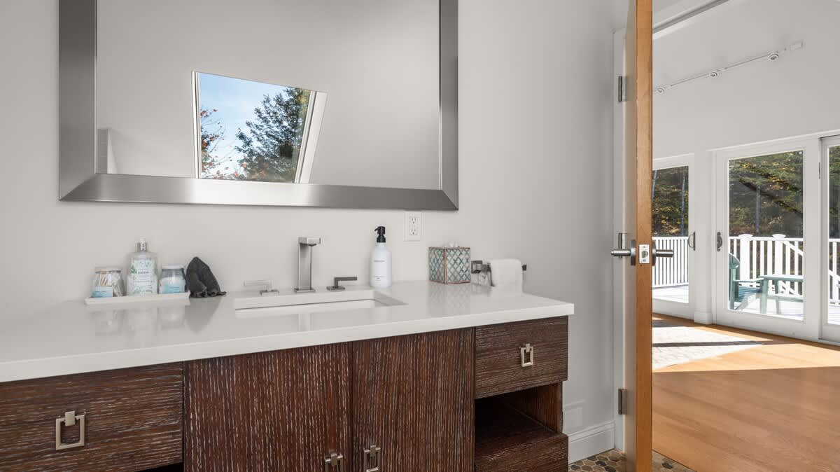 Bathroom vanity with a dark wood cabinet, white countertop, and modern faucet, beneath a large rectangular mirror. The reflection shows an outdoor deck with white railings and trees, accessible through a nearby glass door.