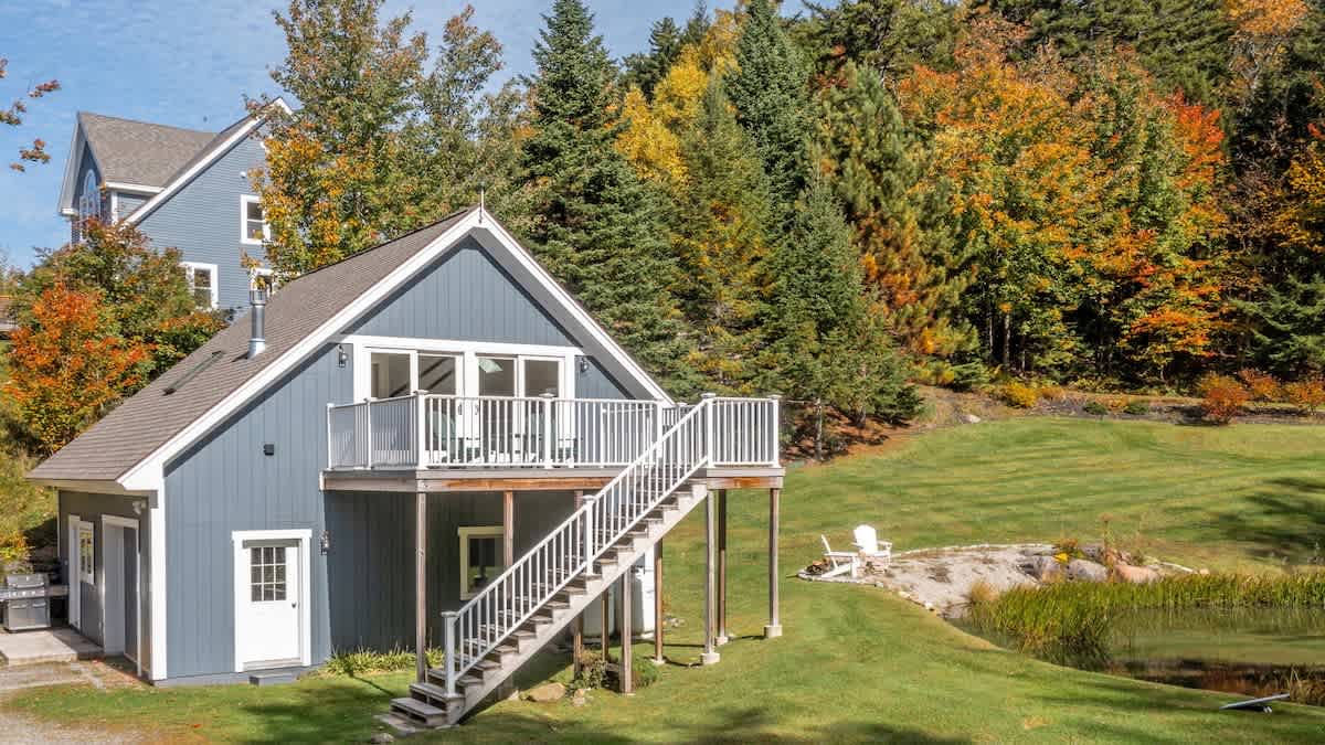 A blue-grey cottage features a raised deck with white railings and stairs, overlooking a grassy lawn and a small pond. The background is filled with vibrant autumn trees and a glimpse of another house.