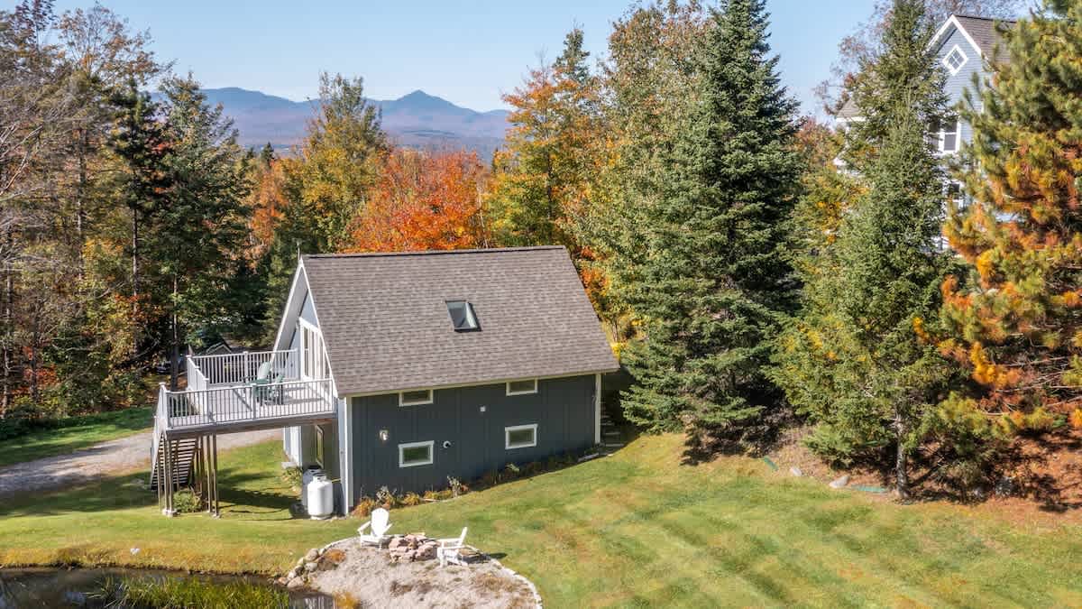 An aerial view captures the blue-grey guest house with a dark roof and skylight, featuring a deck with outdoor seating. A lush green lawn, a small pond, and vibrant autumn trees frame the property, with distant mountains visible on the horizon.