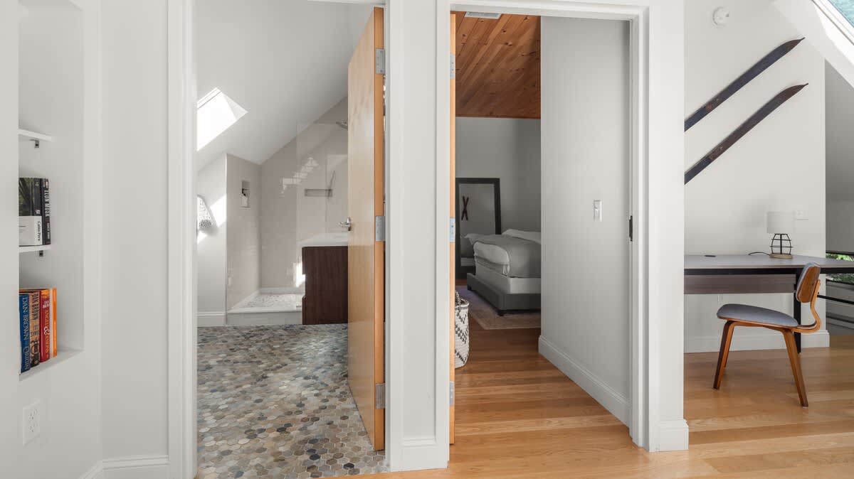 A bright hallway connects the bathroom, visible through an open door with a pebble floor and glass shower, to the bedroom/office area on the right with hardwood floors. Built-in shelving with books is on the left.