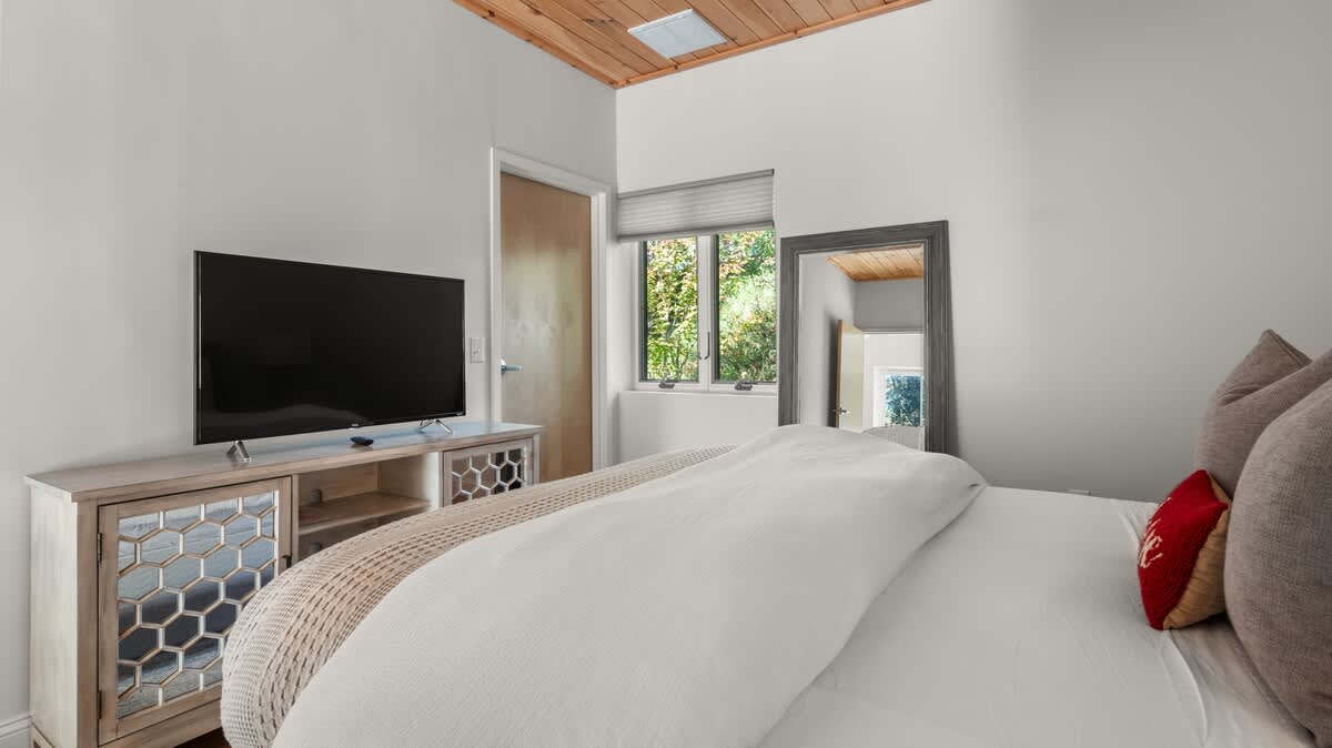 View of the bedroom highlighting the large flat-screen TV on a light wood media console with geometric patterned doors. The queen bed with its gray headboard and white bedding is partially visible, with a wooden ceiling above.