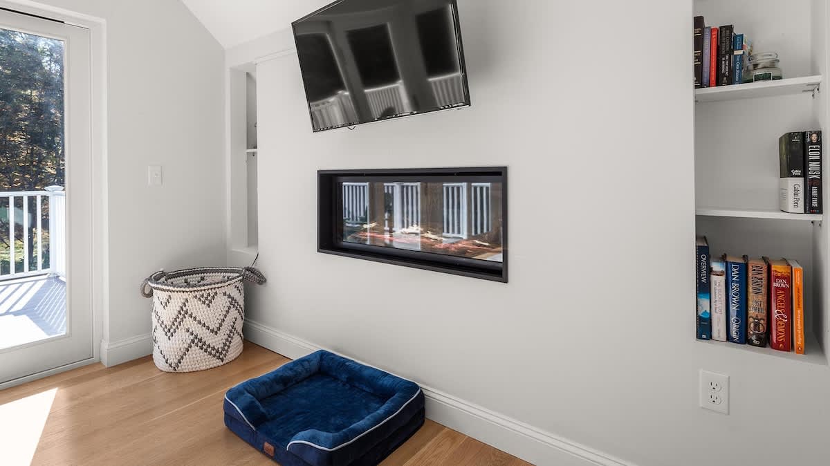A detailed view of the living room's entertainment wall, featuring a wall-mounted flat-screen TV above a sleek electric fireplace. Built-in shelving filled with books flanks the fireplace, and a blue dog bed rests on the light hardwood floor.
