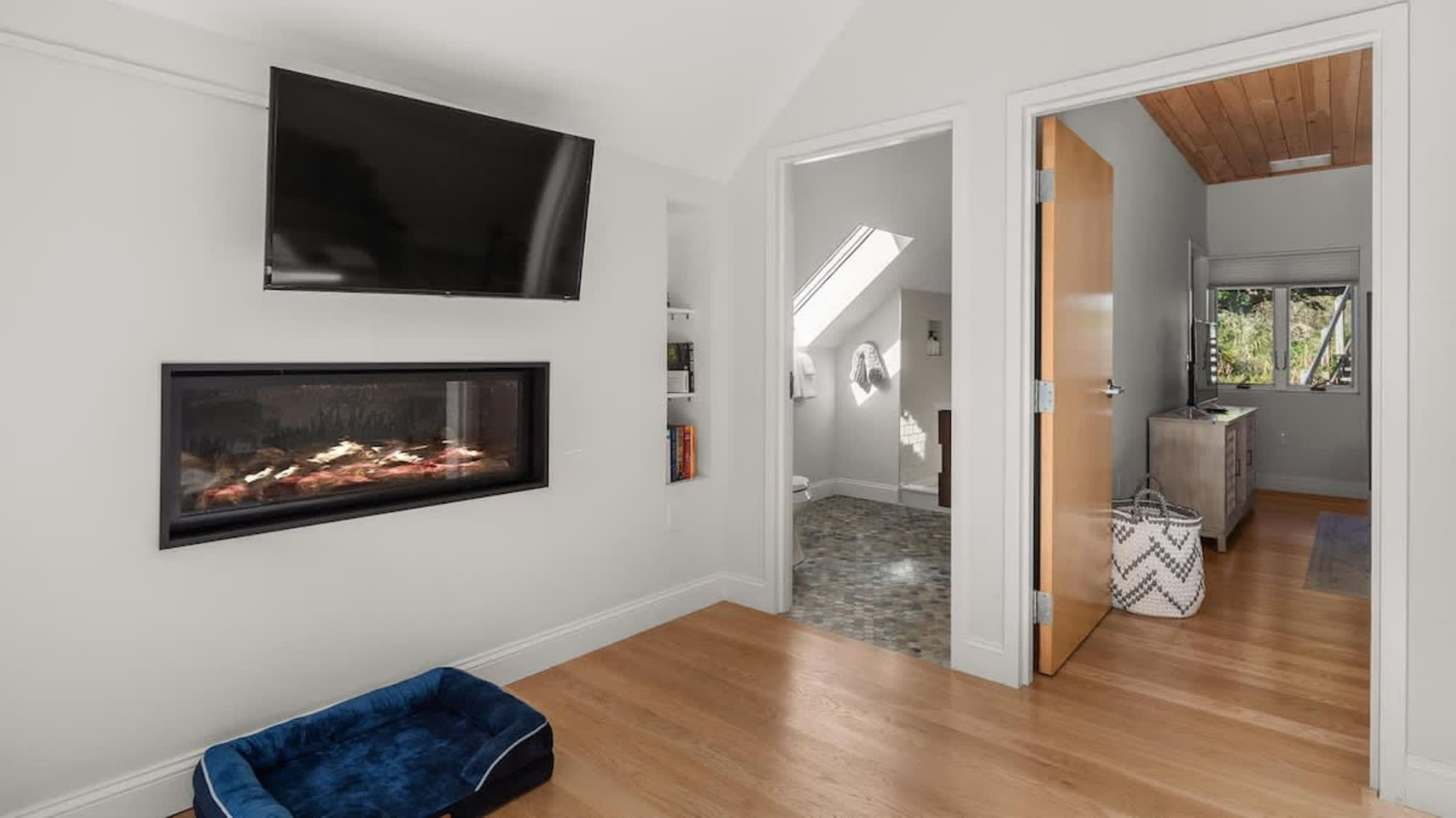 A modern electric fireplace is integrated into a light gray wall, topped by a mounted flat-screen TV. To the right, built-in shelves display books, while a blue dog bed rests on the light hardwood floor. Two open doorways lead to adjacent rooms.