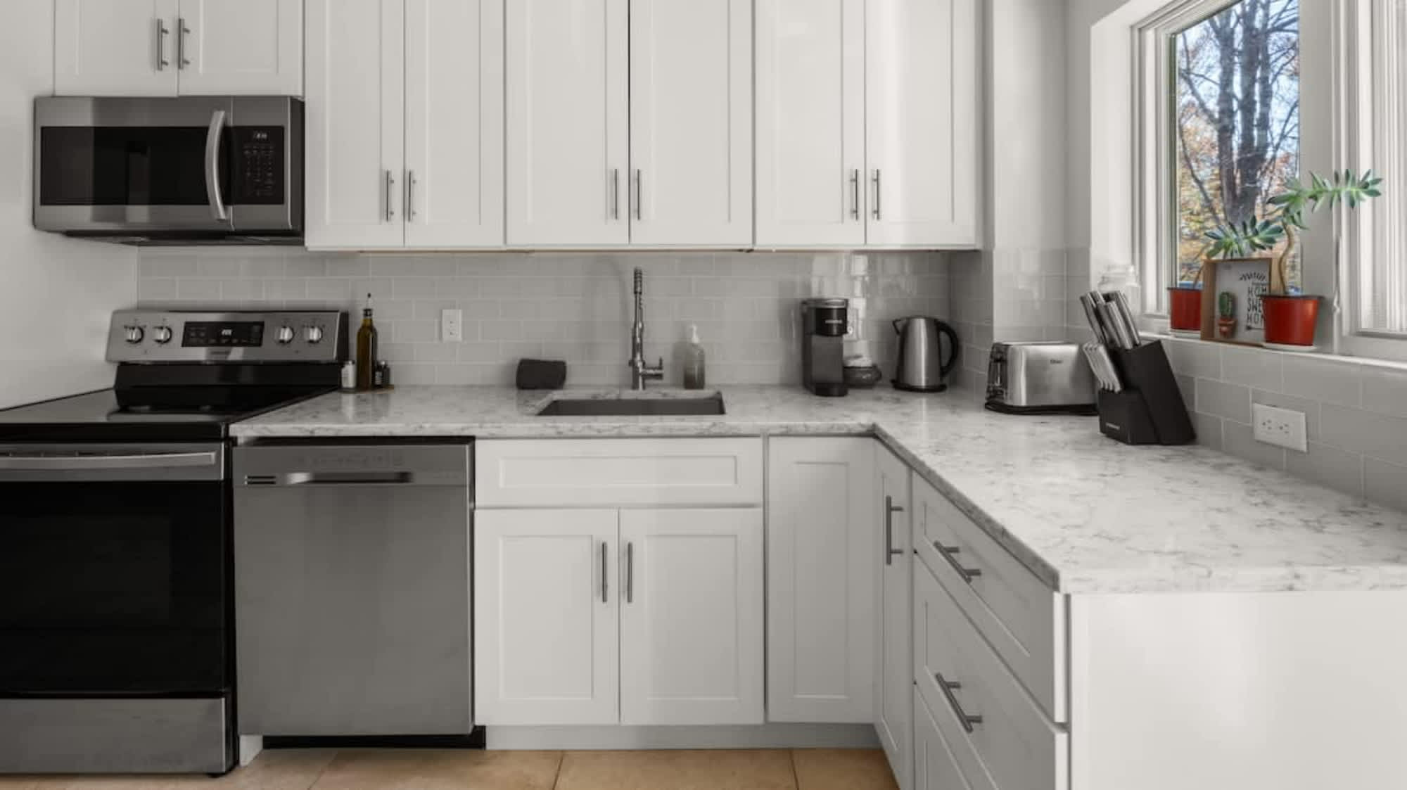 Close-up of the modern kitchen's L-shaped counter, showcasing white shaker cabinets, marble-look countertops, and a subway tile backsplash. Stainless steel appliances like the range, dishwasher, and sink are visible, along with a coffee maker and toaster.