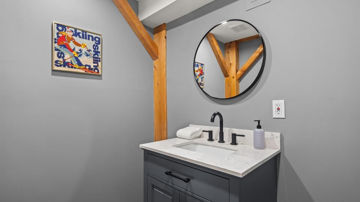 Modern bathroom with a dark gray vanity, white countertop, black faucet, and a round black-framed mirror. A vintage skiing poster adds a pop of color, and exposed wooden beams are visible in the corner.