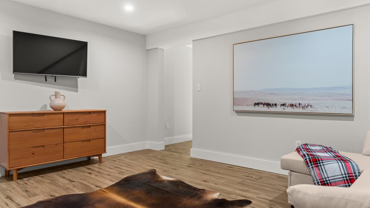 Spacious living area featuring a wall-mounted flat-screen TV above a mid-century modern wooden dresser. A large landscape artwork hangs on an adjacent wall, and a dark animal hide rug is on the light wood floor.