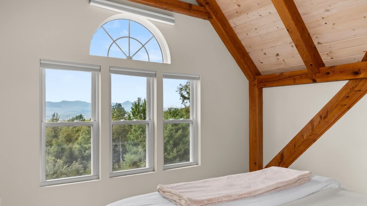 Bright bedroom with a large window array, including an arched transom window, offering expansive views of green trees and distant mountains. A bed with a light blanket is positioned below the windows.