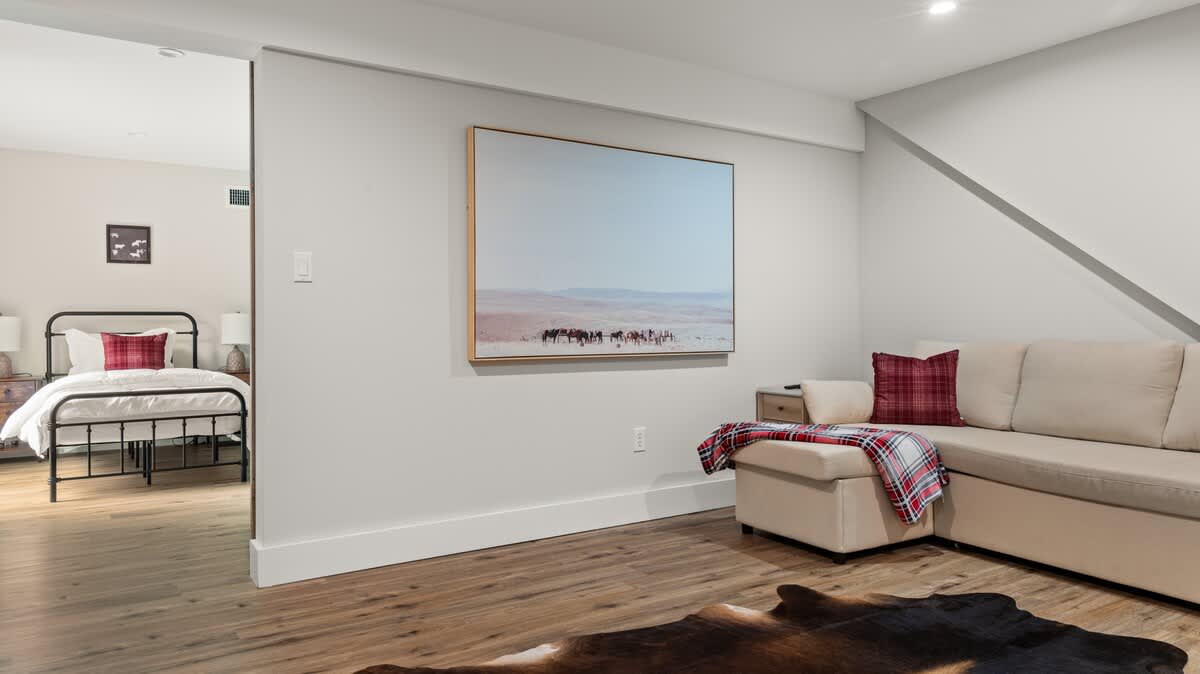 Cozy living area with a light beige sectional sofa, a red plaid throw blanket, and a large landscape artwork featuring horses on a desert plain. A bedroom with two beds is visible through an opening in the wall.