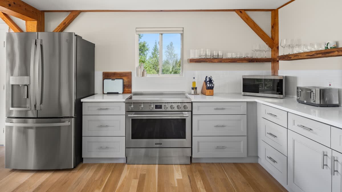 Modern kitchen with light gray shaker cabinets, white quartz countertops, stainless steel appliances including a large refrigerator and range, and exposed wooden beams overhead. A window above the sink offers a view of trees.