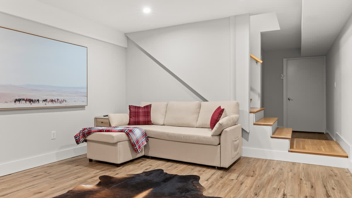 Inviting living area with a light beige sectional sofa, a red plaid throw, and a large landscape artwork. Wooden stairs with a handrail lead up, and a dark animal hide rug covers part of the light wood floor.