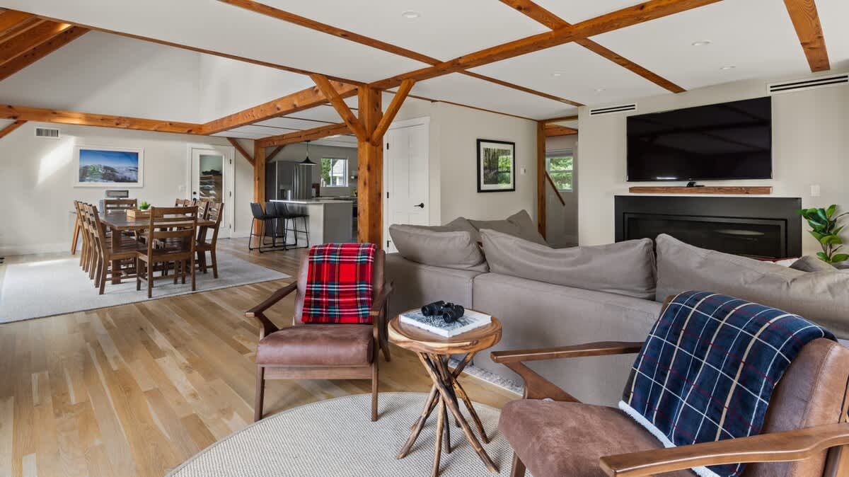 Spacious open-concept living and dining area with light hardwood floors, exposed wooden beams, a large sectional sofa, two armchairs, and a long wooden dining table with chairs. A large TV is mounted above a fireplace.